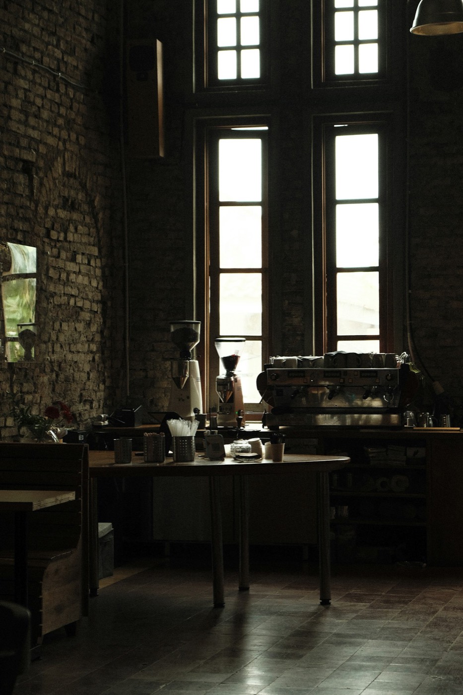 Barra de café con máquina de espresso y molinos frente a ventanales altos, en un local de ladrillo.