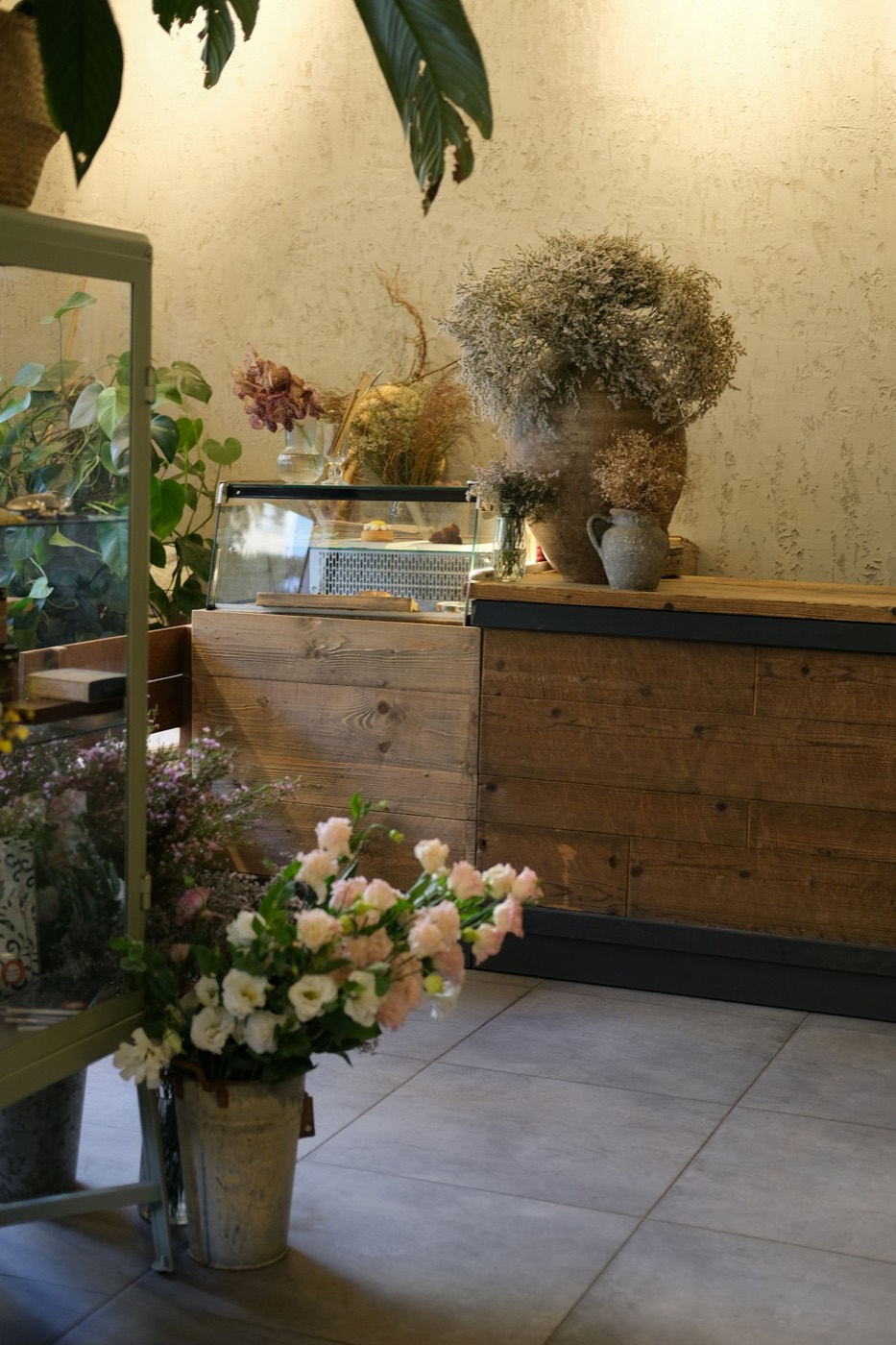 Mostrador de madera con vitrina, flores secas y un balde de flores frescas en primer plano.